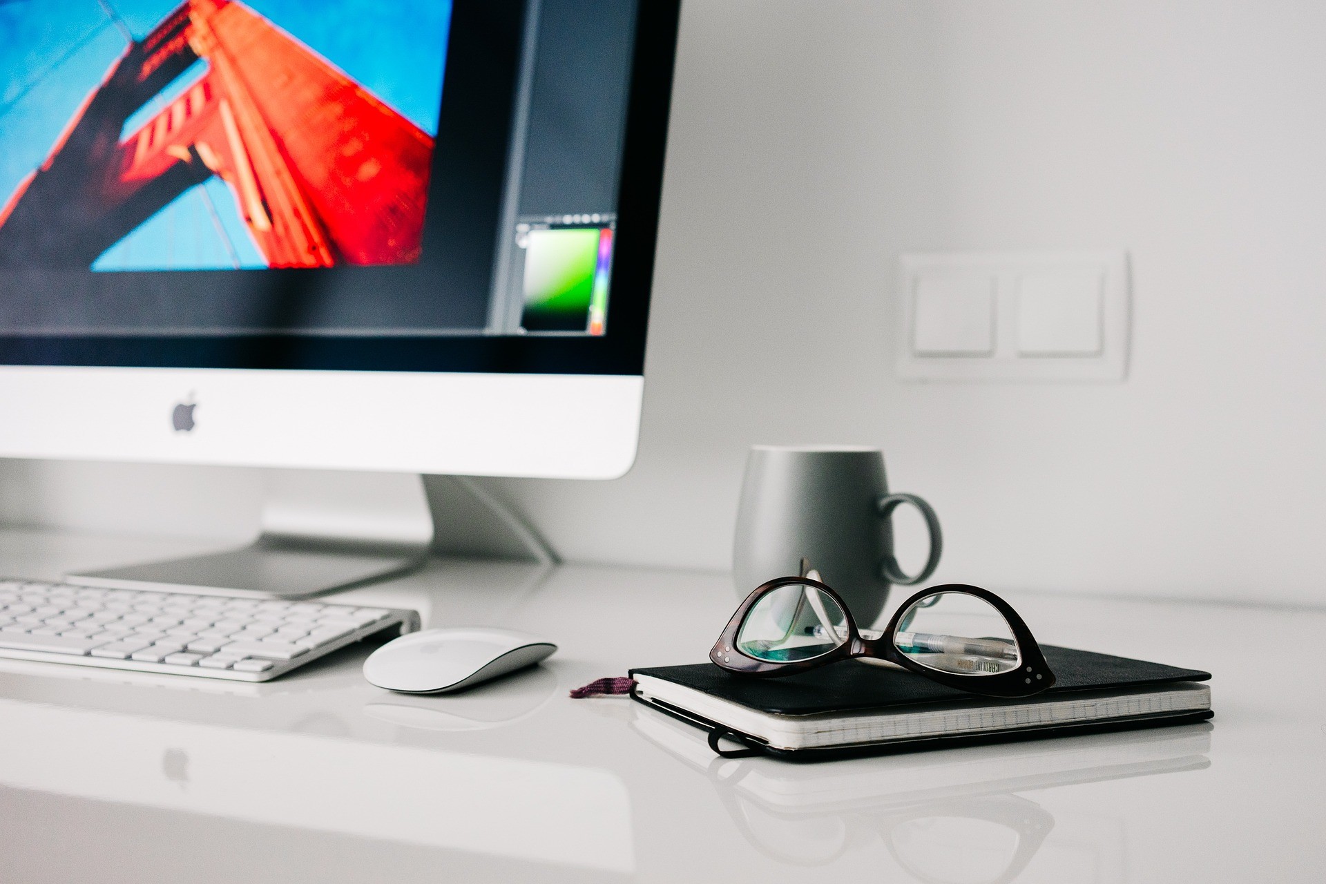A desk with an iMac, coffee mug and a small notebook with some spectacles on it.